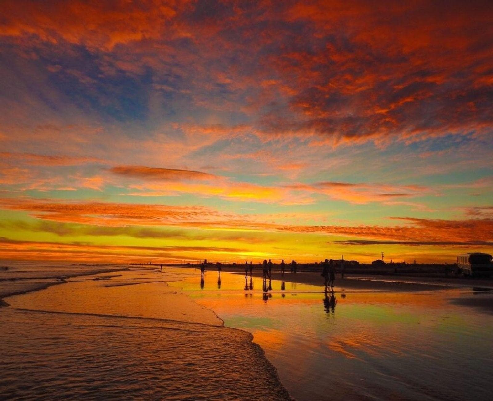 Pessoas caminhando pela areia molhada da Praia do Cassino ao entardecer, com o céu refletido na superfície.
